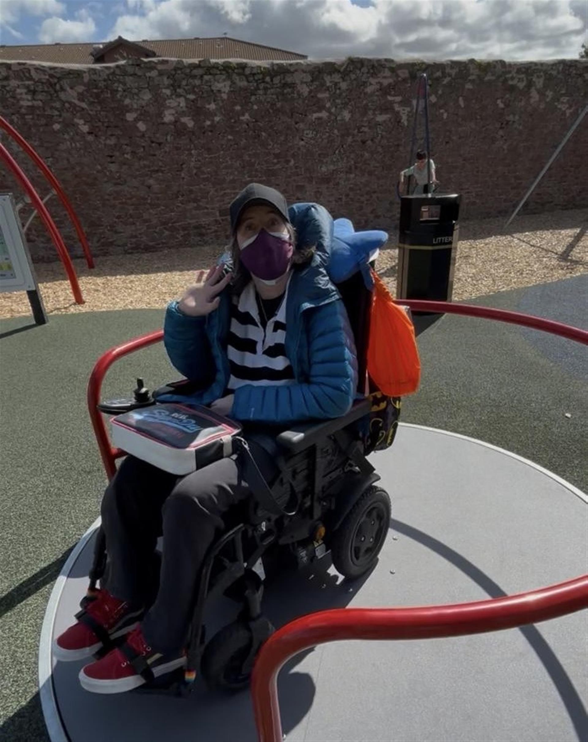 Euan's Guide Ambassador Karine sitting in her wheelchair, facing the camera with a mask on and waving, having fun on a wheelchair accessible roundabout