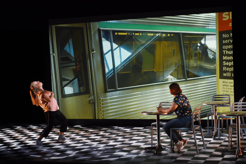 Julia sitting at a table set on a checkerboard floor with another actor standing
