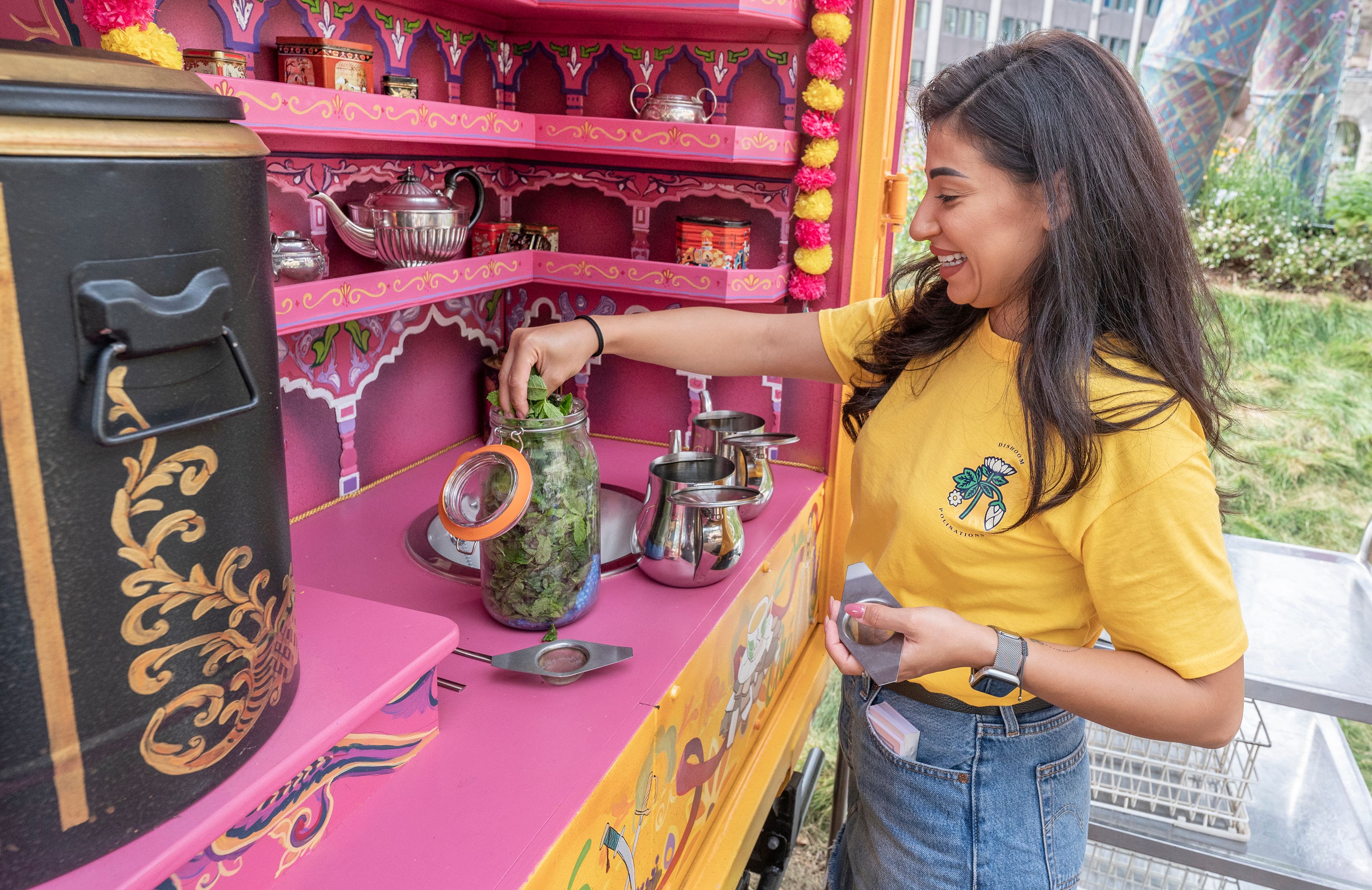 A woman in a yellow t shirt and jeans stands at a pink stall as she takes mint tea leaves out of a glass jar