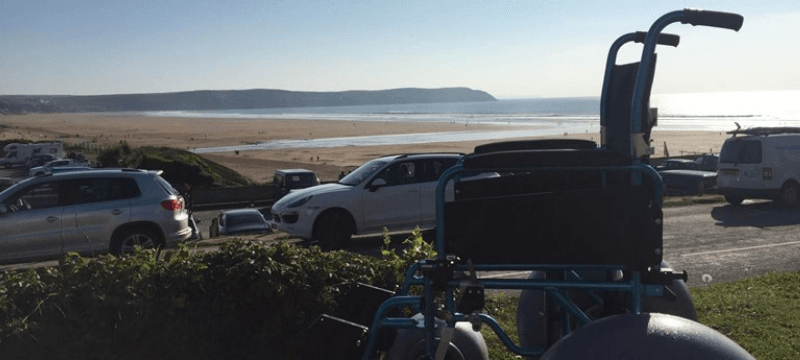 Image of a beach wheelchair sitting on a grass embankment in front of a road with cars and the beach behind them.